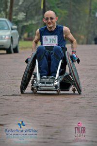 A front shot of Jayden in his racing wheelchair on a cobblestone street wearing a racing bib number 724 at the Tyler Azalea race.