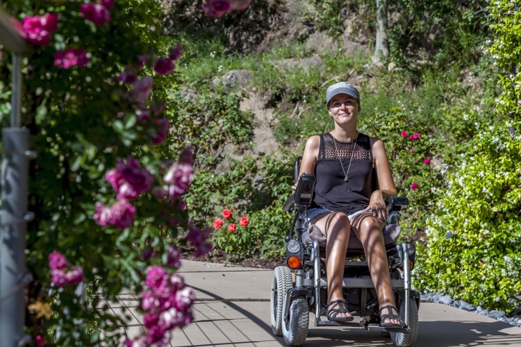 Bibi in her wheelchair on a cement path with greenery and flowers surrounding her.