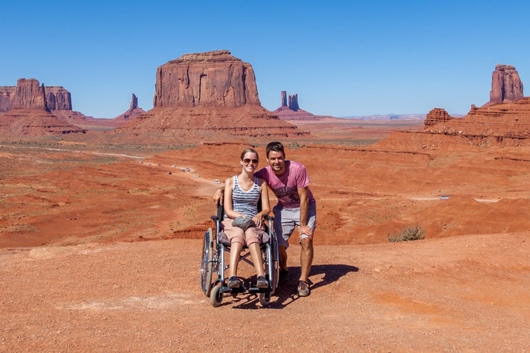 Bibi in a wheelchair and her boyfriend standing by her side in Monument Valley Navajo Tribal Park with rock formations in the background.