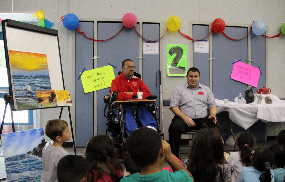 Man in wheelchair with another man in front of children in classroom