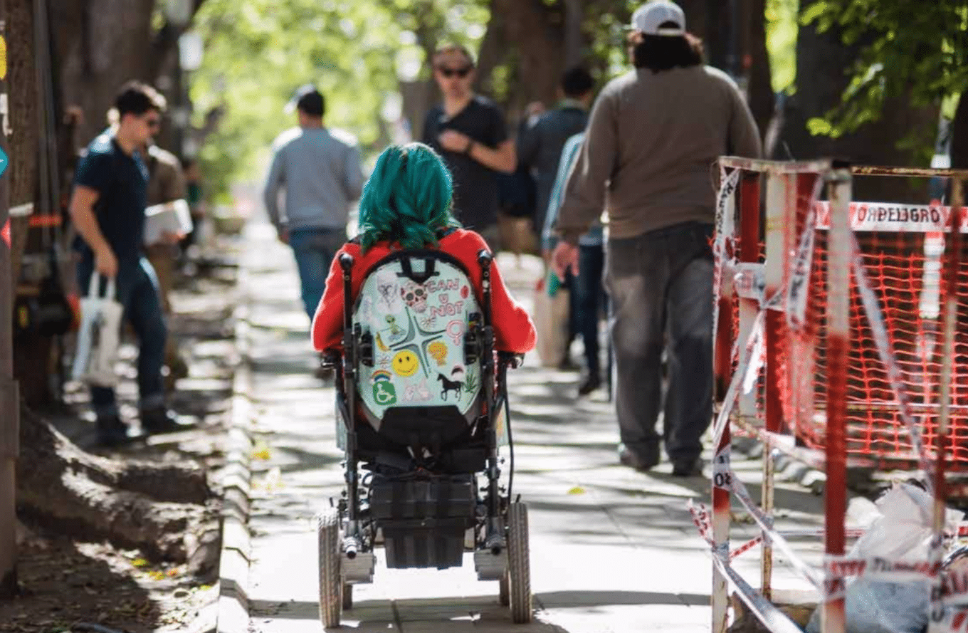 Juana, a young woman with blue hair in her power wheelchair rolling down a city sidewalk