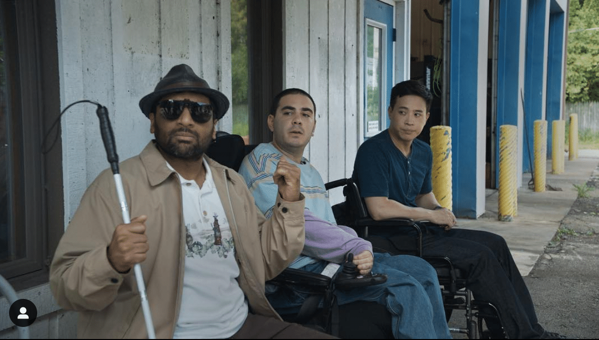 Visually impaired man, a power wheelchair using man, and a manual wheelchair using man sitting against the outside wall of a service station