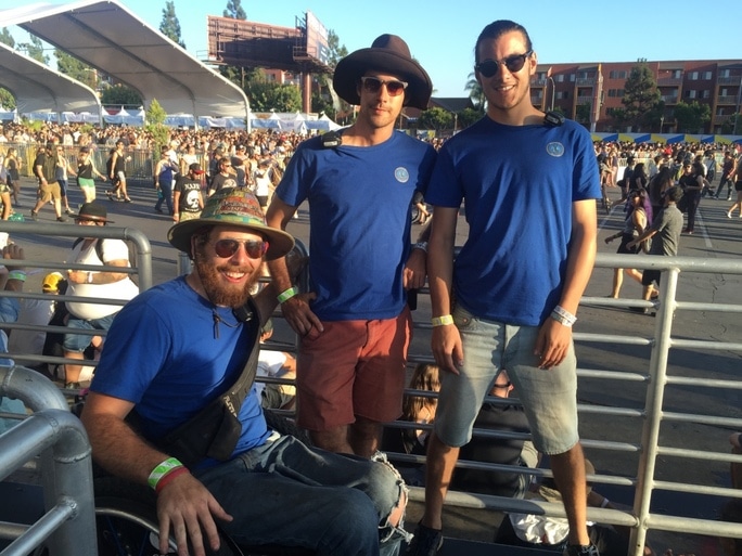 Three Accessible Festivals staff members in blue shirts, one in a wheelchair, two standing at an outdoor event venue