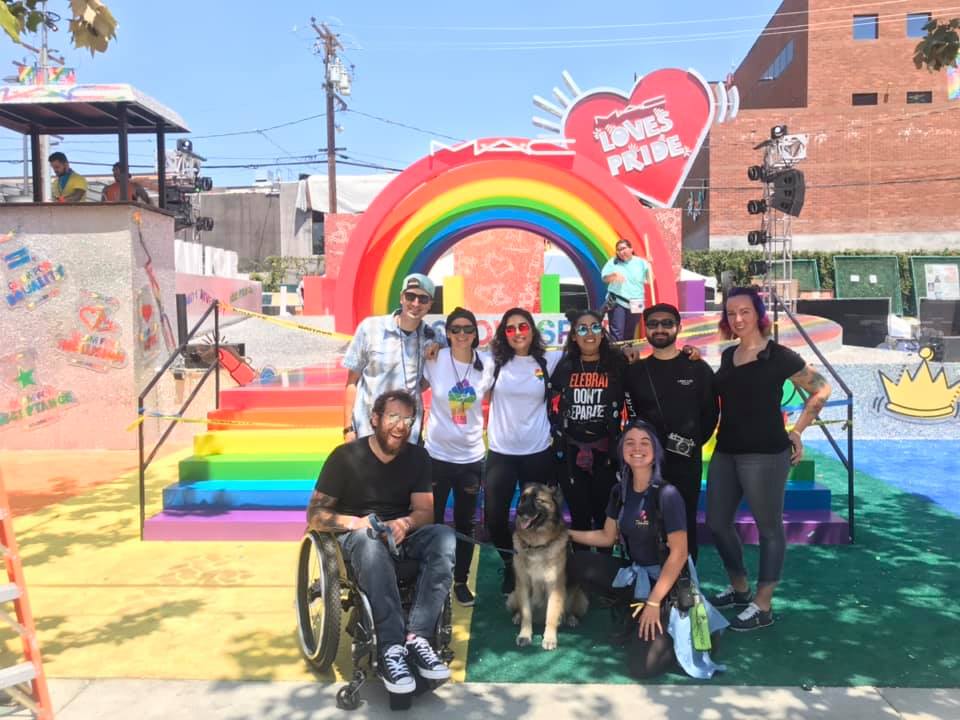 Group of people, one in wheelchair and one with a service dog, in front of a rainbow display with a "MAC Loves Pride" sign. 