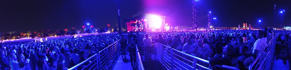 Nighttime festival with crowd of people. We see a ramp in the middle of the crowd and a lit up stage in the distance. 