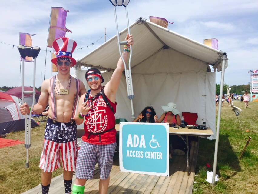 Two men standing, one with a cast on his leg holding up a crutch in front of the ADA Access Center tent at a festival