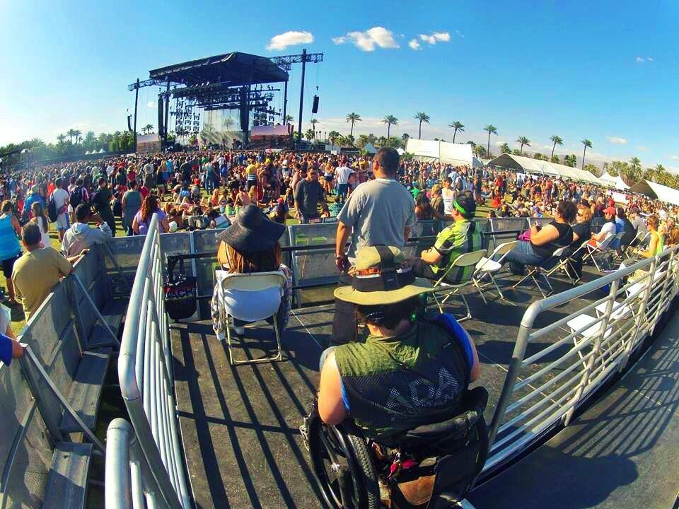A crowd at an outdoor festival. One man is in a wheelchair in the accessible seating area with an ADA assistant shirt on.