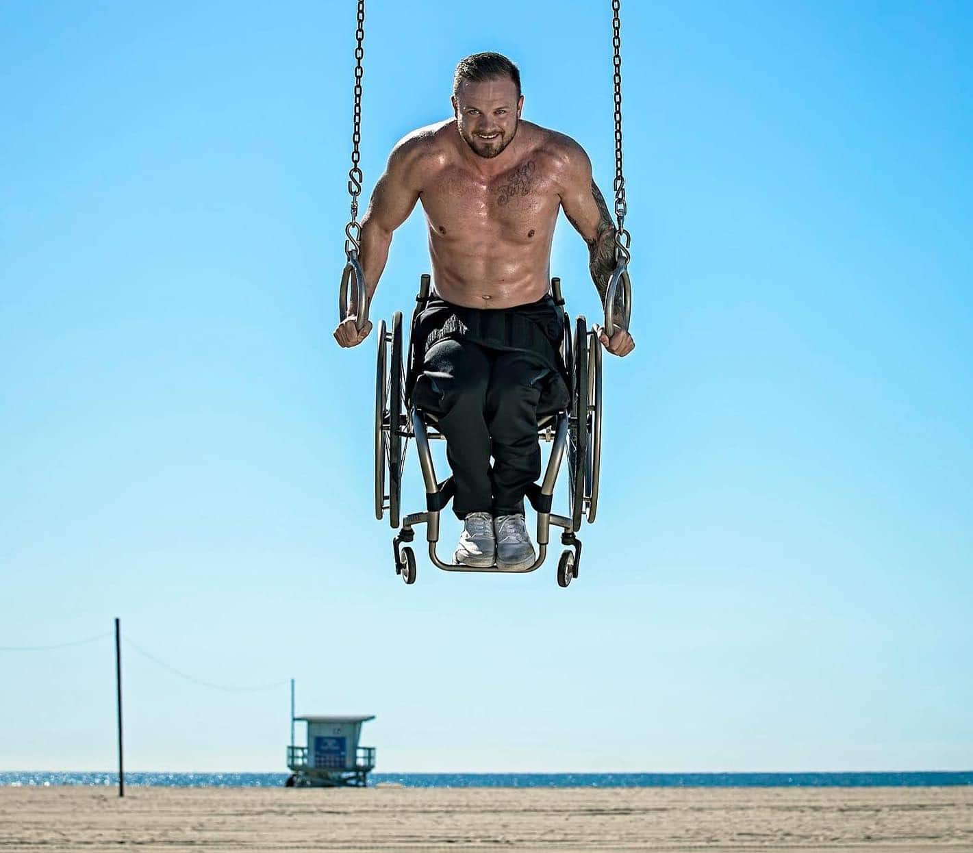 Richard Corbett is shirtless with black pants sitting in his wheelchair while doing a gymnastics ring hold at a beach
