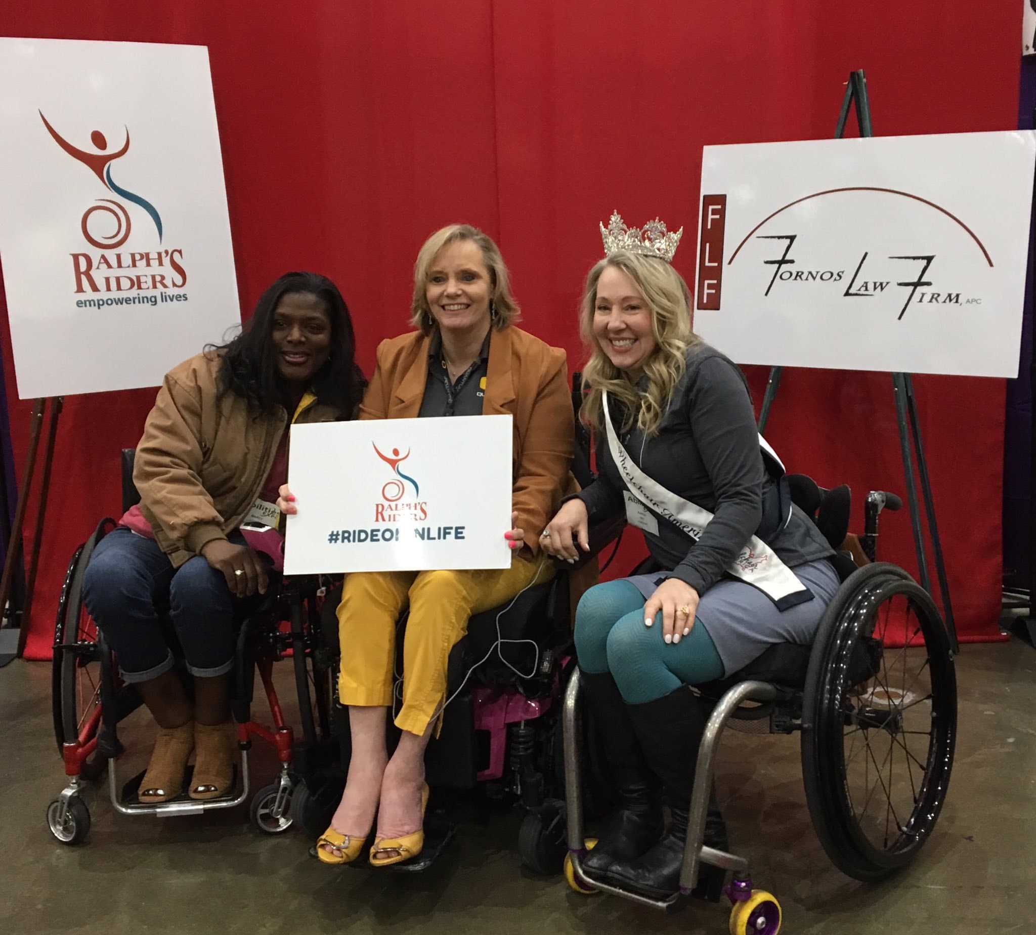 Three women in wheelchairs in front of Ralph's Riders and Fornos Law signs. One is holding a #rideoninlife sign.