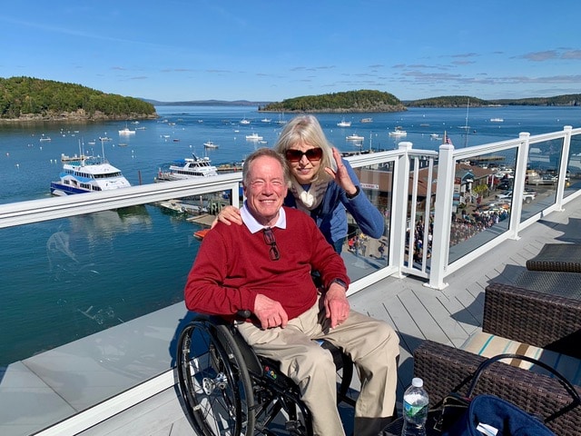 Jim Parsons is sitting in his manual wheelchair wearing a read sweater and khaki pants. His wife is posing next to him for the photo. They are on an elevated area overlooking a harbor with many boats on the water.