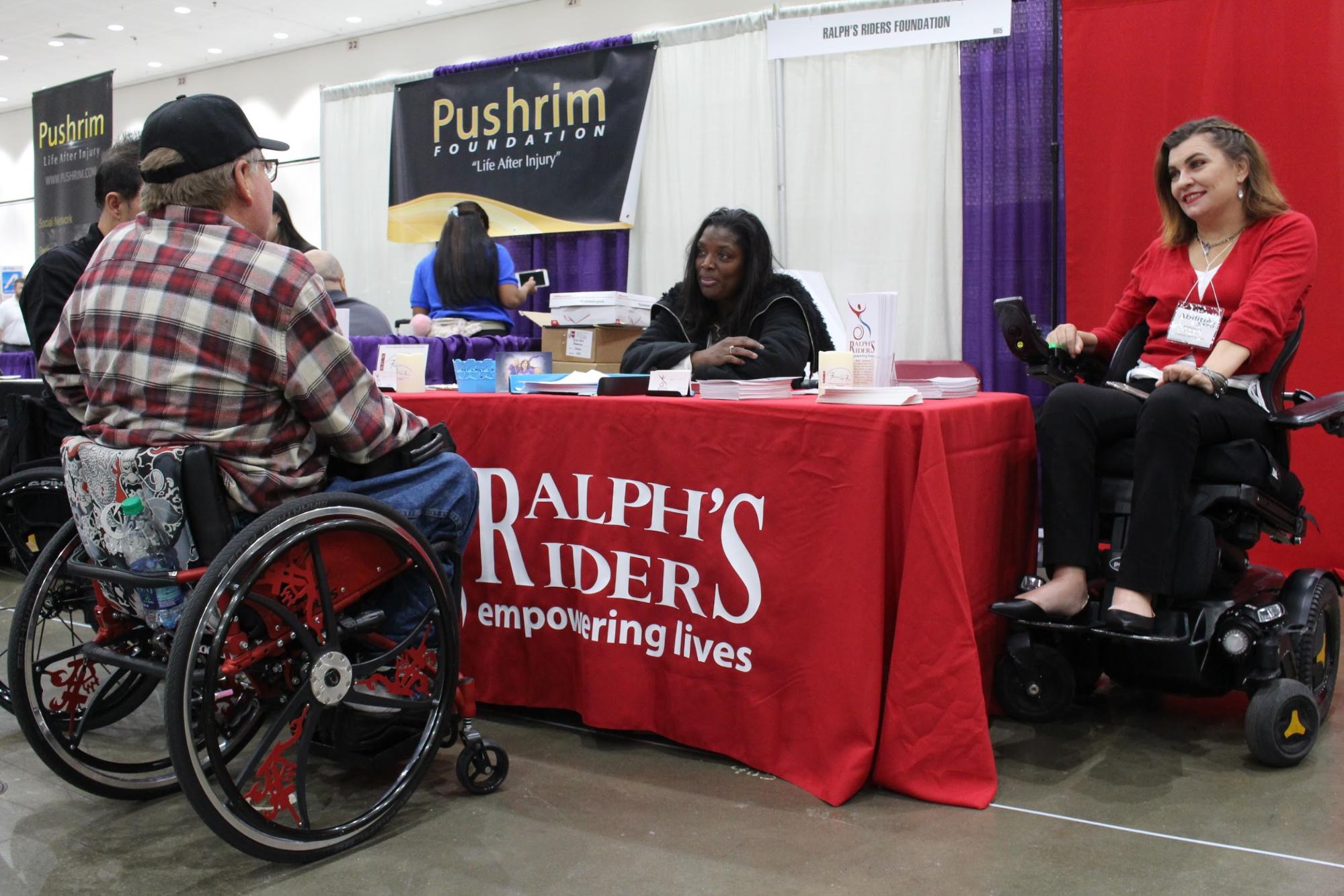 Marcy Lovett and Margarita Elizondo working the Ralph's Riders booth at Abilities Expo. A man in a wheelchair is visiting the booth.