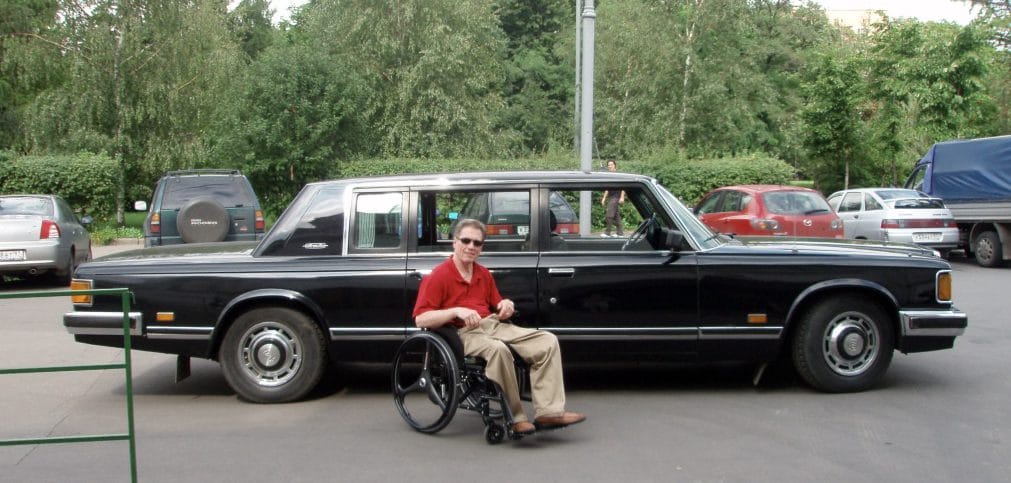 Jim Parsons is sitting in his manual wheelchair wearing a red polo shirt and khaki pants. He is in front of a long black limo-like car from the 70s or 80s.