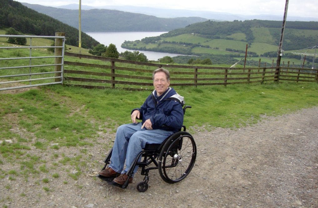 Jim Parsons is sitting in his wheelchair in a blue jacket. In the background, we see grassy hills and a winding river.