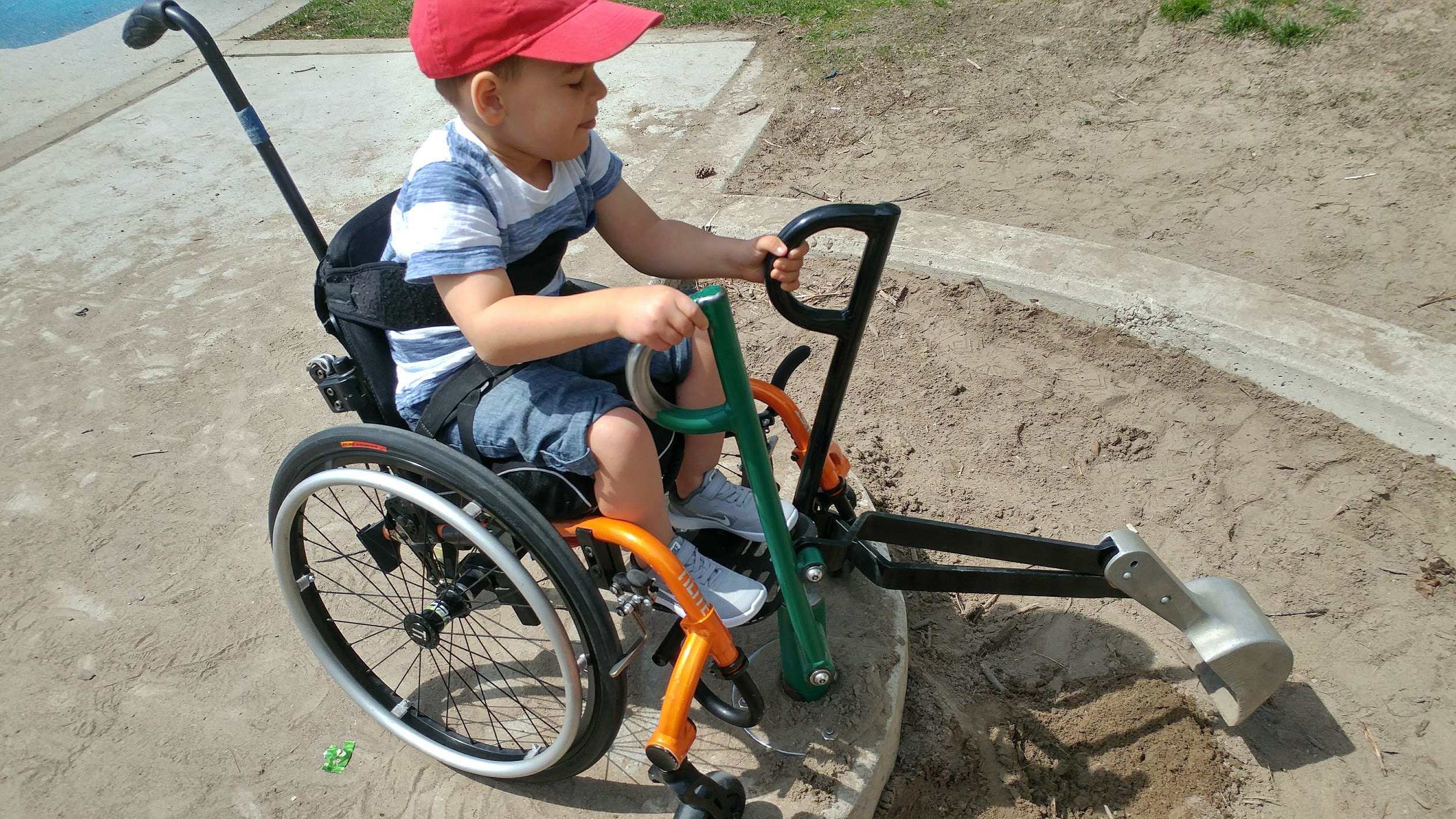 a little boy with a red baseball cap is sitting in his orange wheelchair and using an adaptive tool to dig in the sand