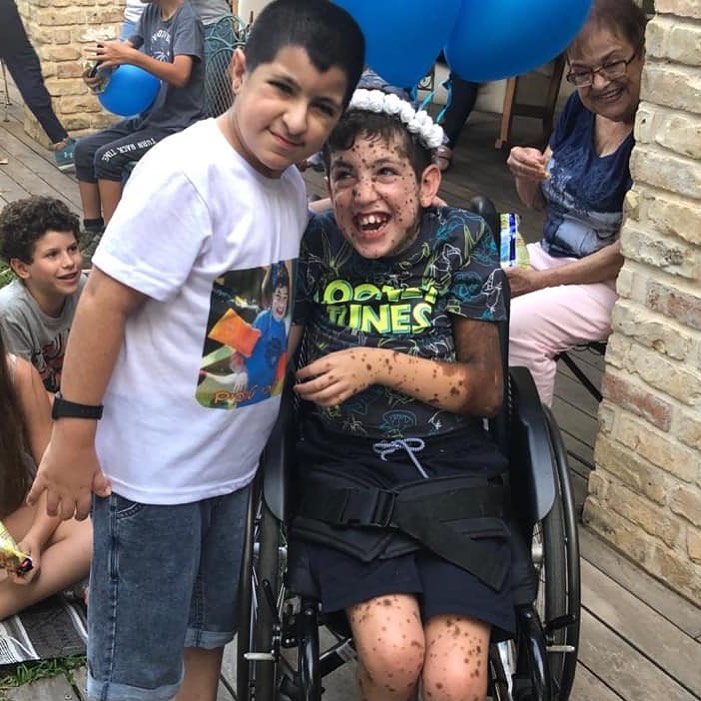 A young boy stands with his arm around Erez who is in his wheelchair. Many children are in the background.