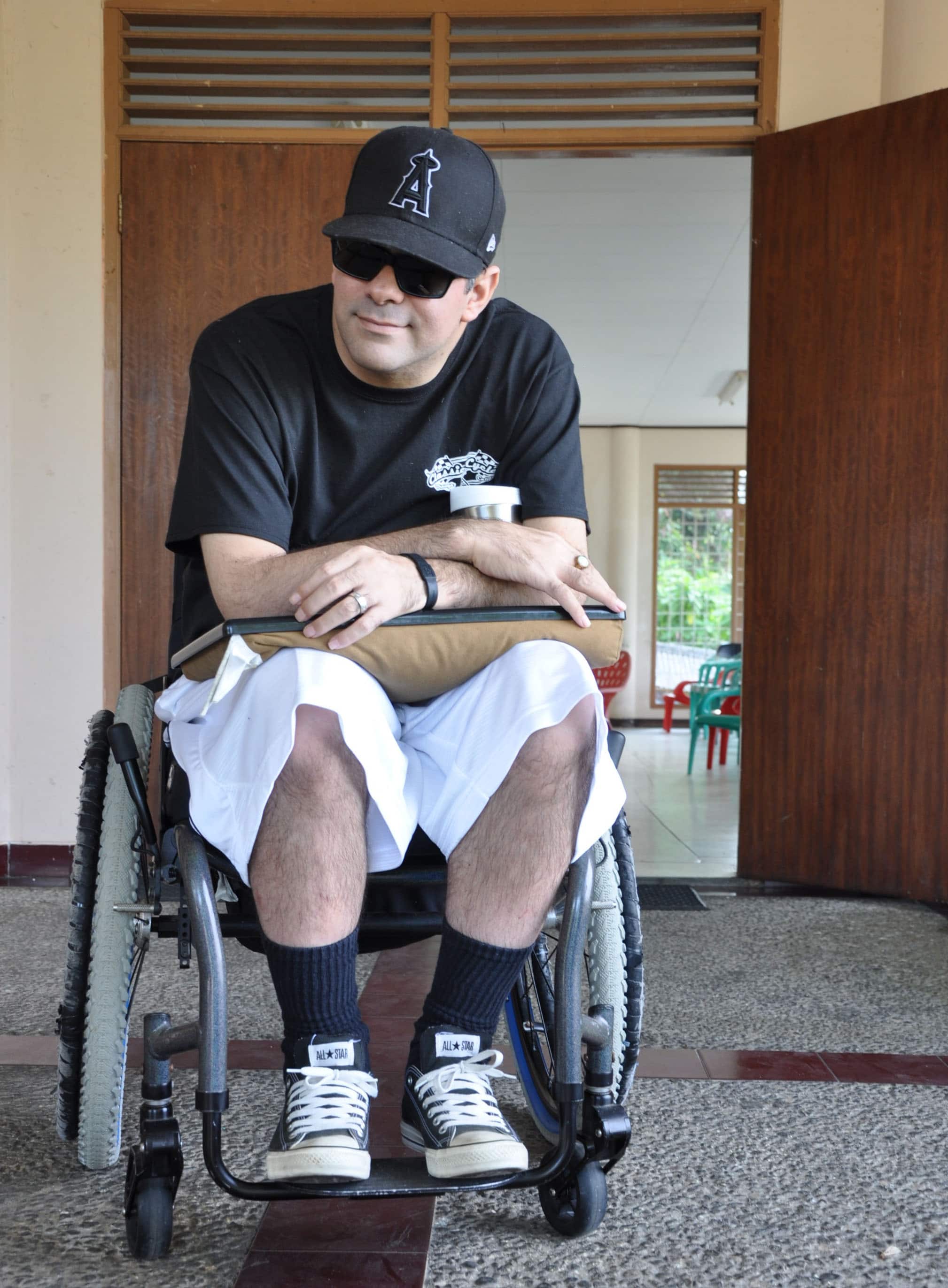 Damien Minna sits in his wheelchair wearing a baseball cap and black sunglasses.