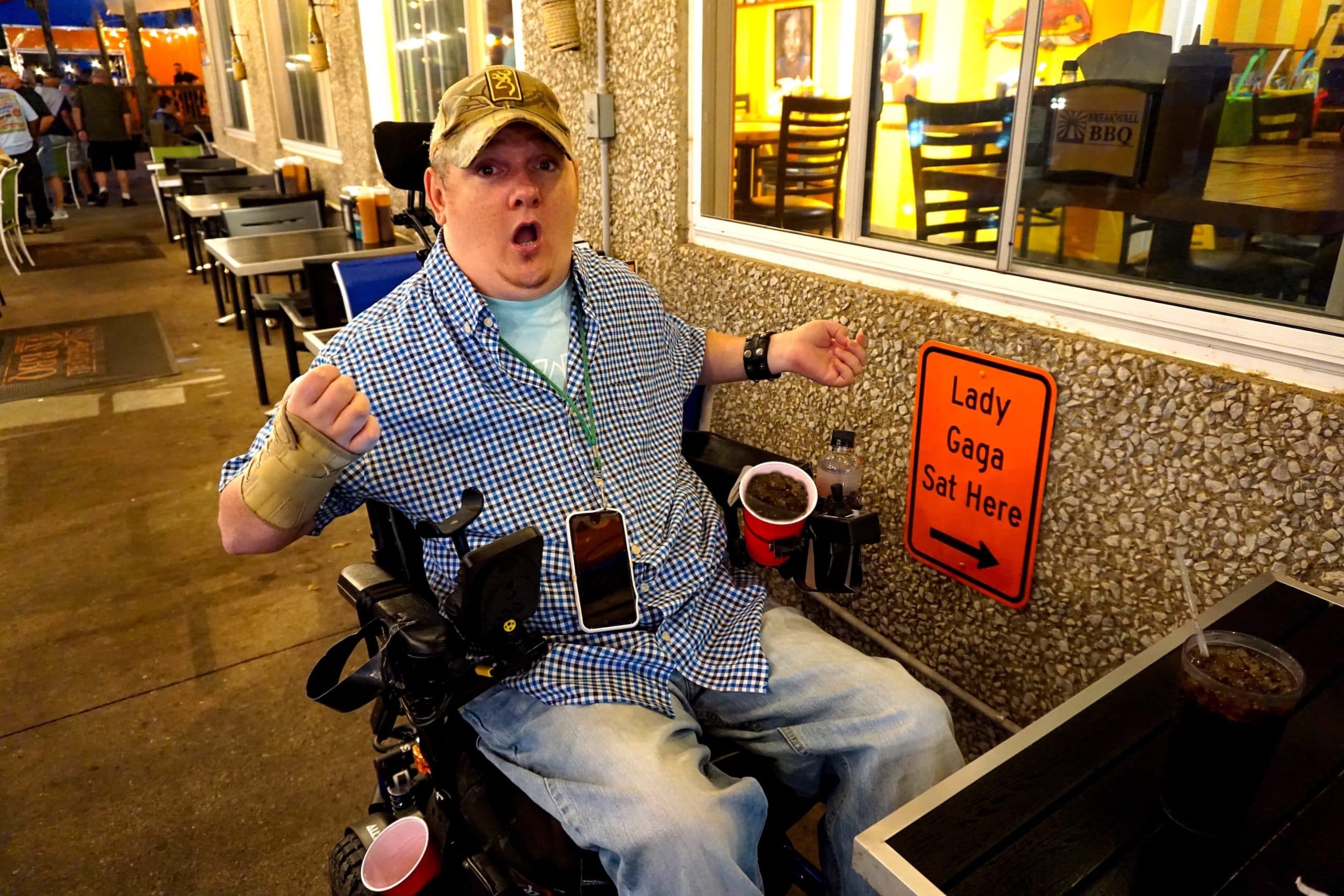 Tim sits at an outdoor table at a restaurant. The sign at his table reads "Lady Gaga sat here."