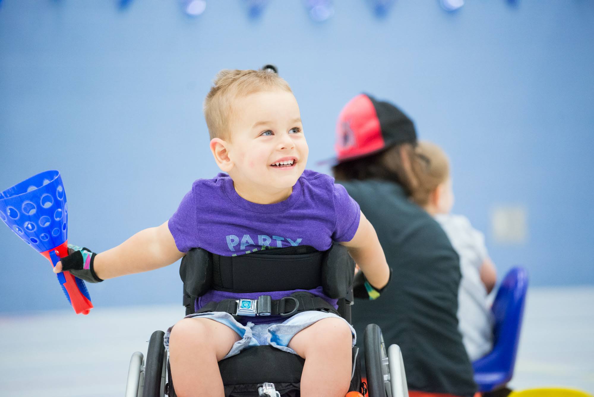 Noah, a little boy, is in his wheelchair in a gym. His has a big smile on his face, and we see some other people in the background. 