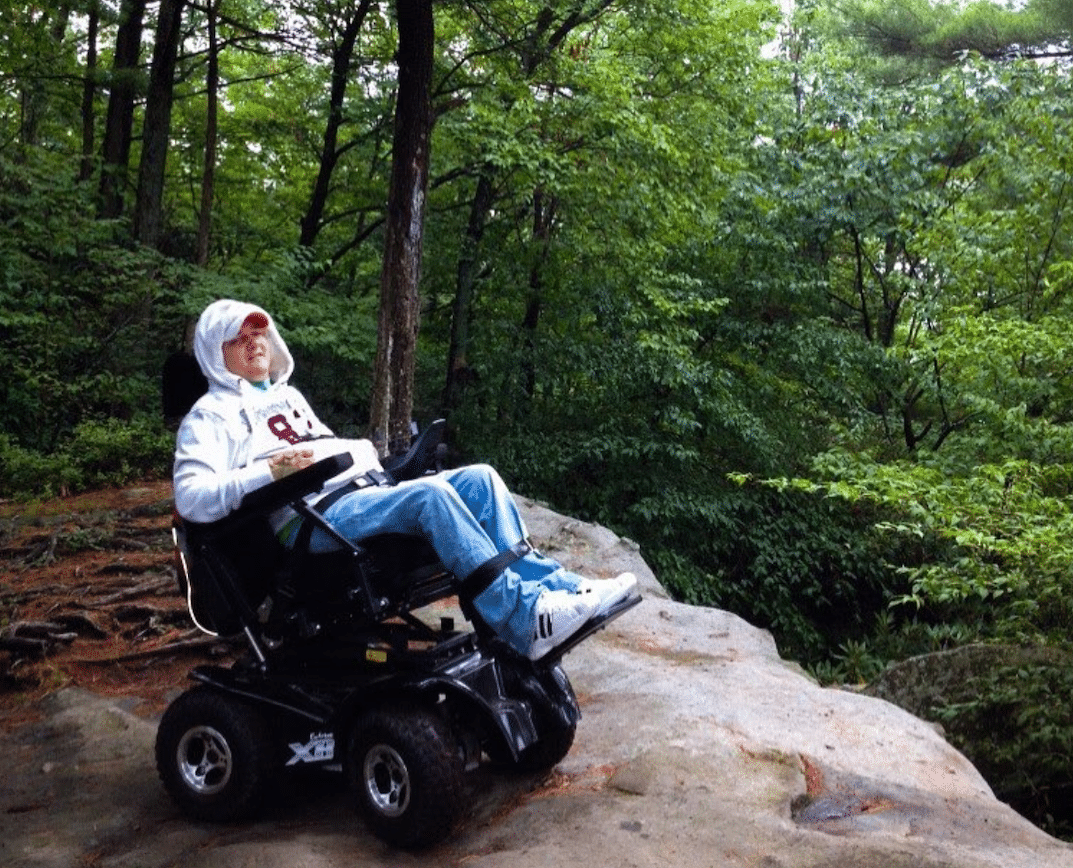 Tim Taylor sits in his power wheelchair on a rocky cliff with green trees in the background.