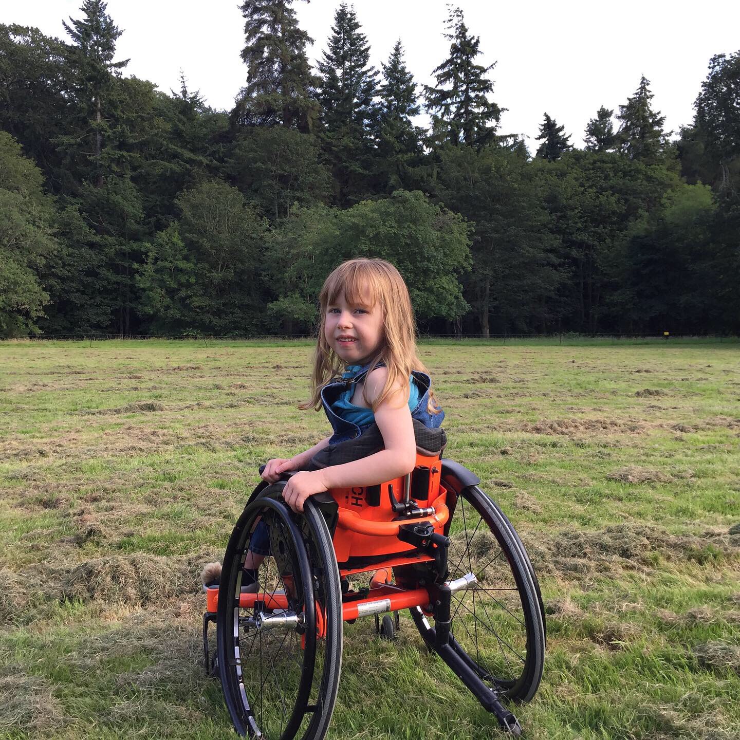 Eudora, a little girl with dirty blond hair, is in a grassy field sitting in her wheelchair looking over her shoulder at the camera.