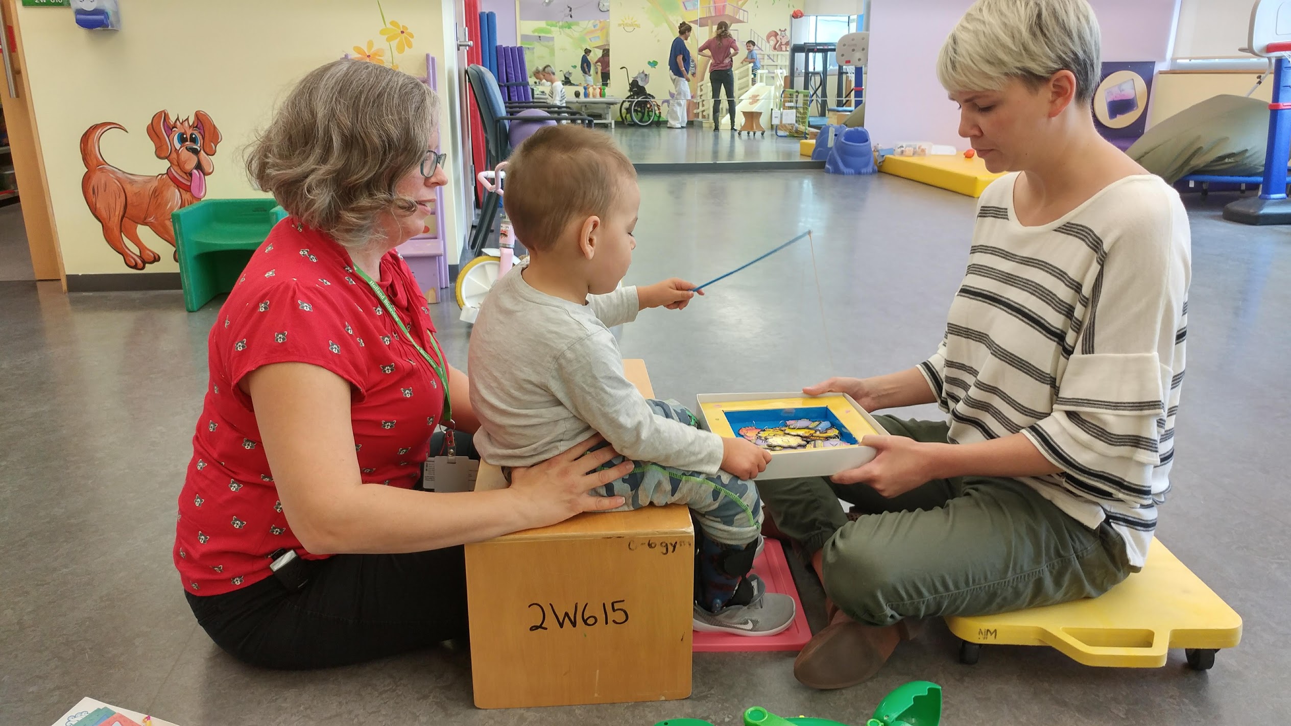 Noah, a little boy, is sitting on a wooden block with a physical therapist supporting his back. Another PT is holding a box while Noah uses a stick to go fishing for items in the box.