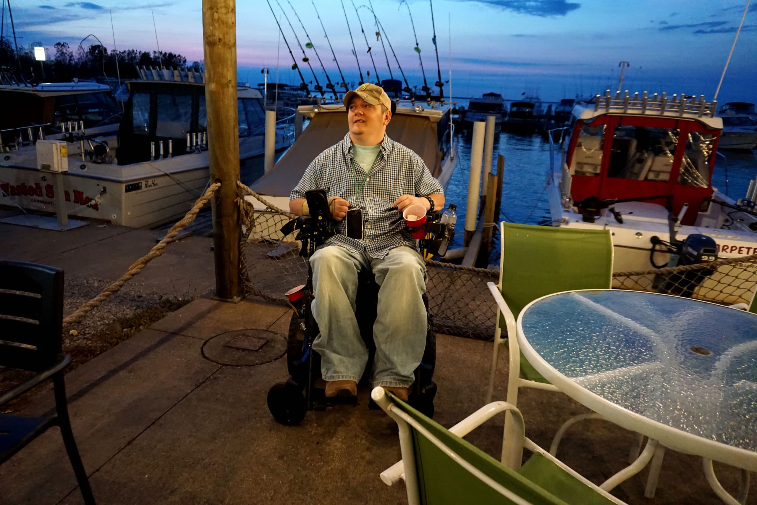 Tim sits in his power wheelchair on a dock. A lake is in the background.