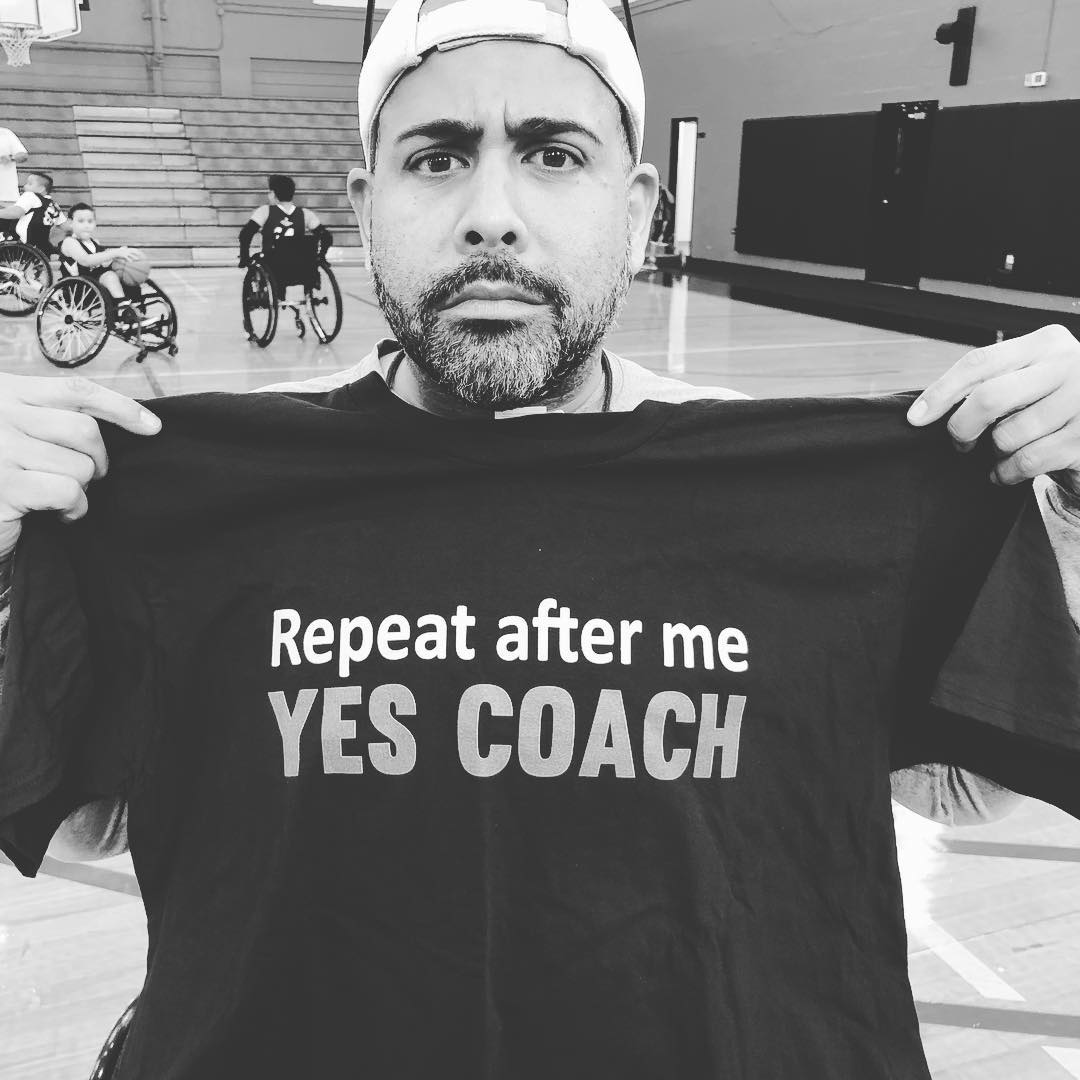 Luis holding up a t-shirt that reads, "Repeat after me: Yes, coach."