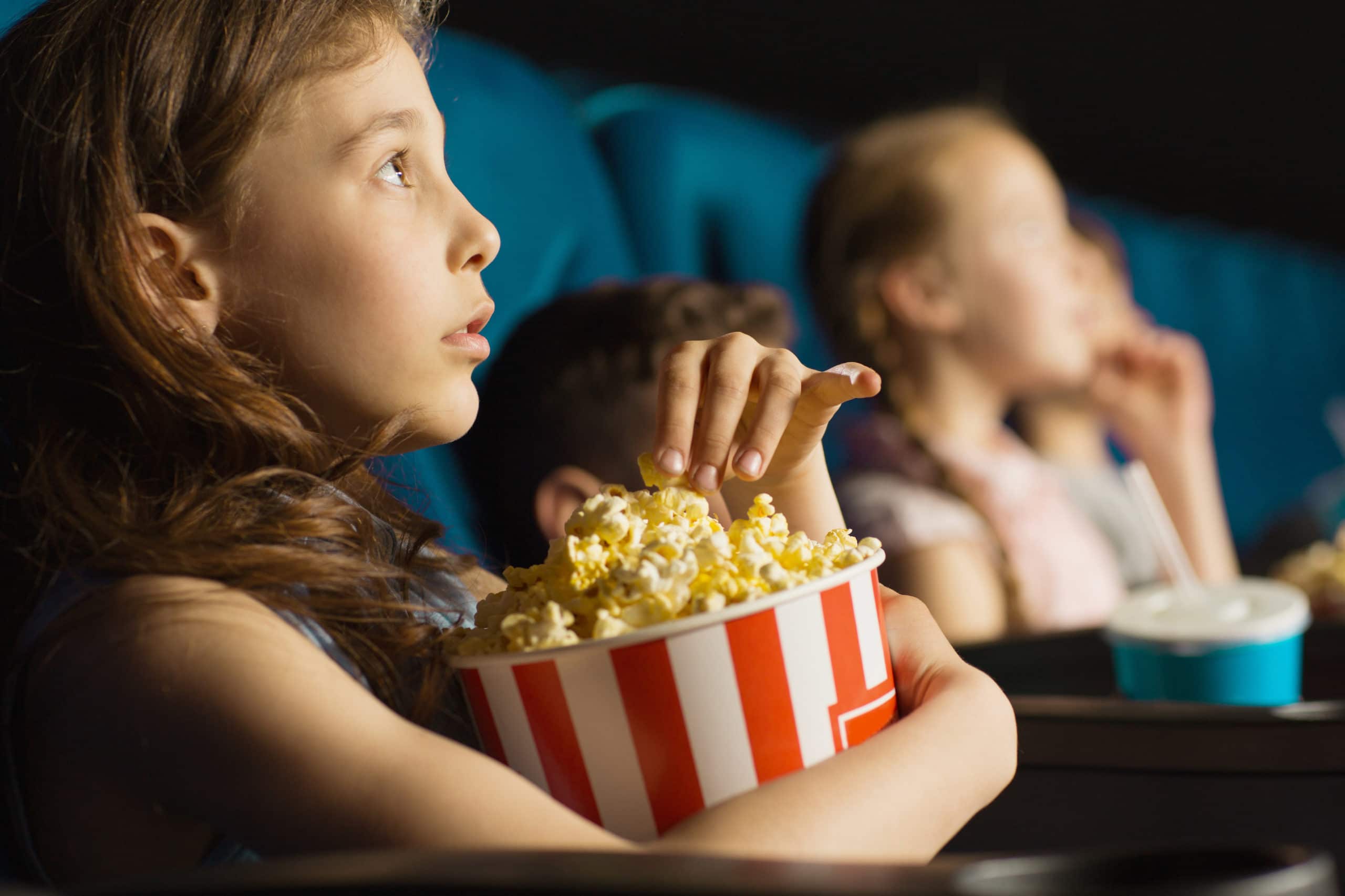 Close up shot of a pretty little girl grabbing popcorn from the bucket while watching a movie at the cinema looking entertained and fascinated.