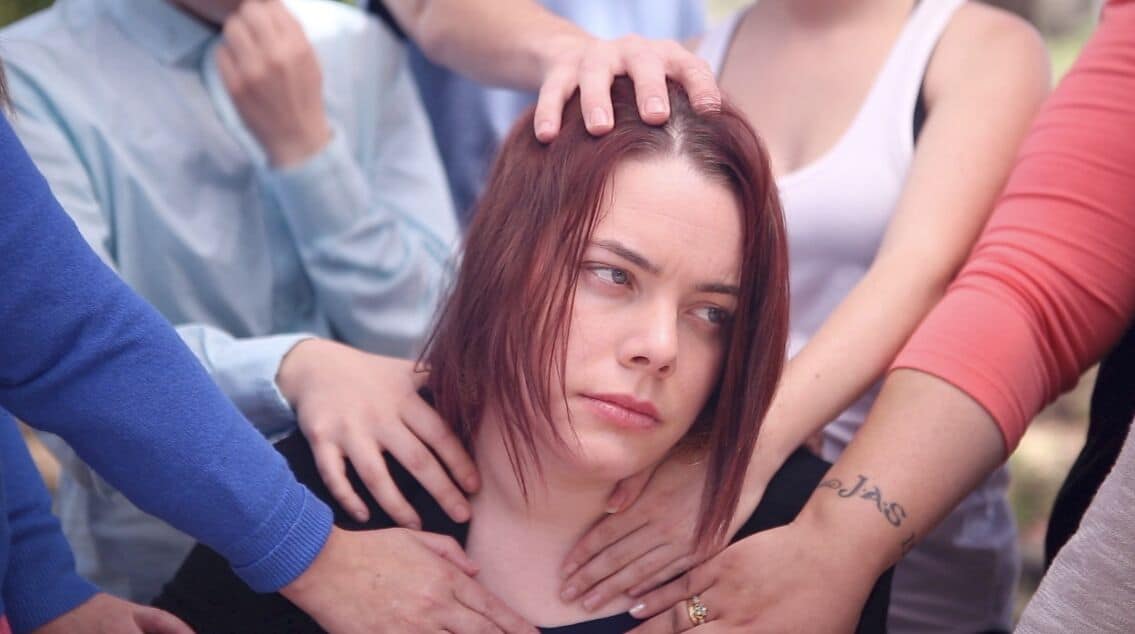 A woman gazing off screen with hands of people whose faces we don't see are touching her head and shoulders.