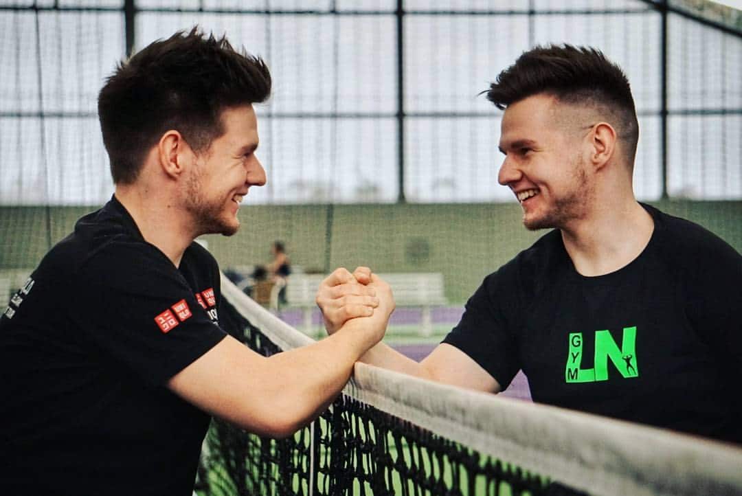 A profile view of twins Max and Marcus smiling and locking hands across a tennis net.