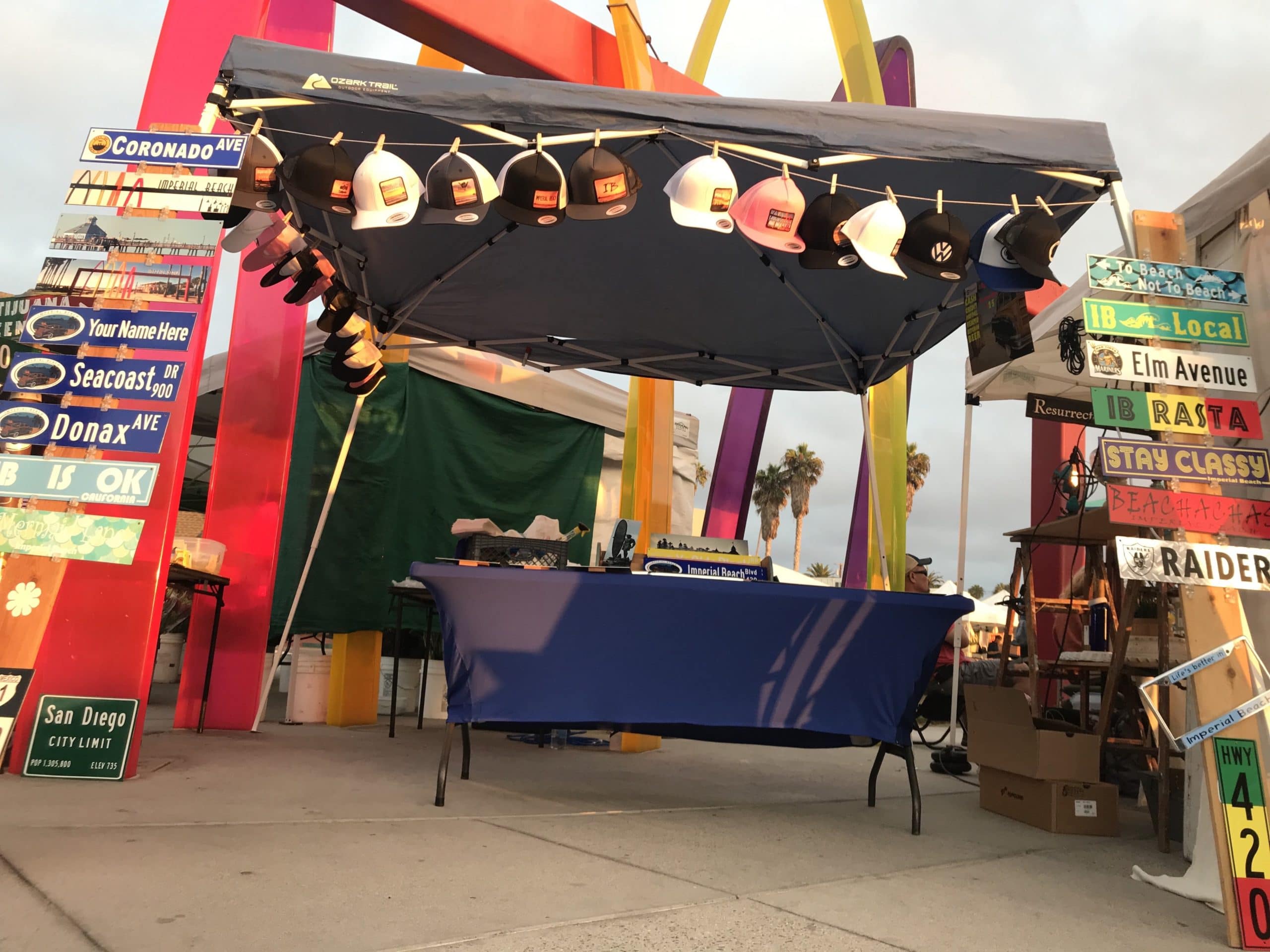 Coronado Sign booth at farmers market with signs and hats for sale.