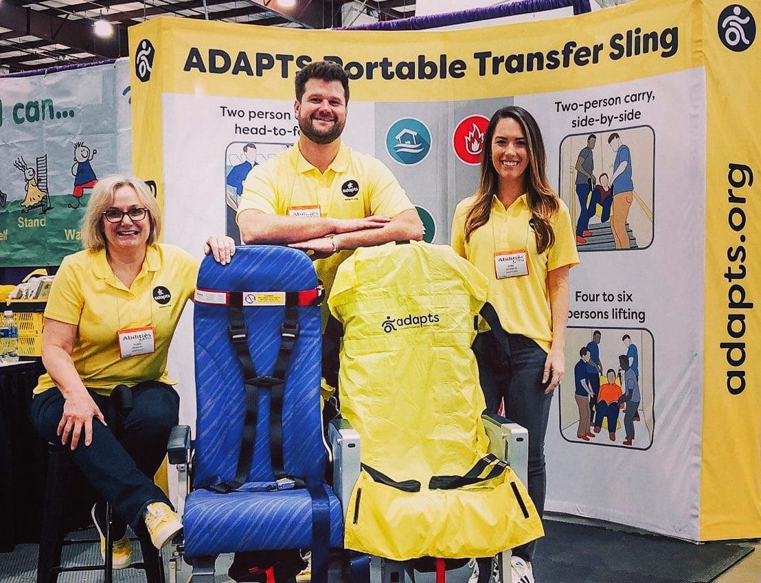 Robin Wearley and a man and woman dressed in yellow ADAPTS t-shirts standing behind airplane seats, one of which has an ADAPTS sling draped over it.