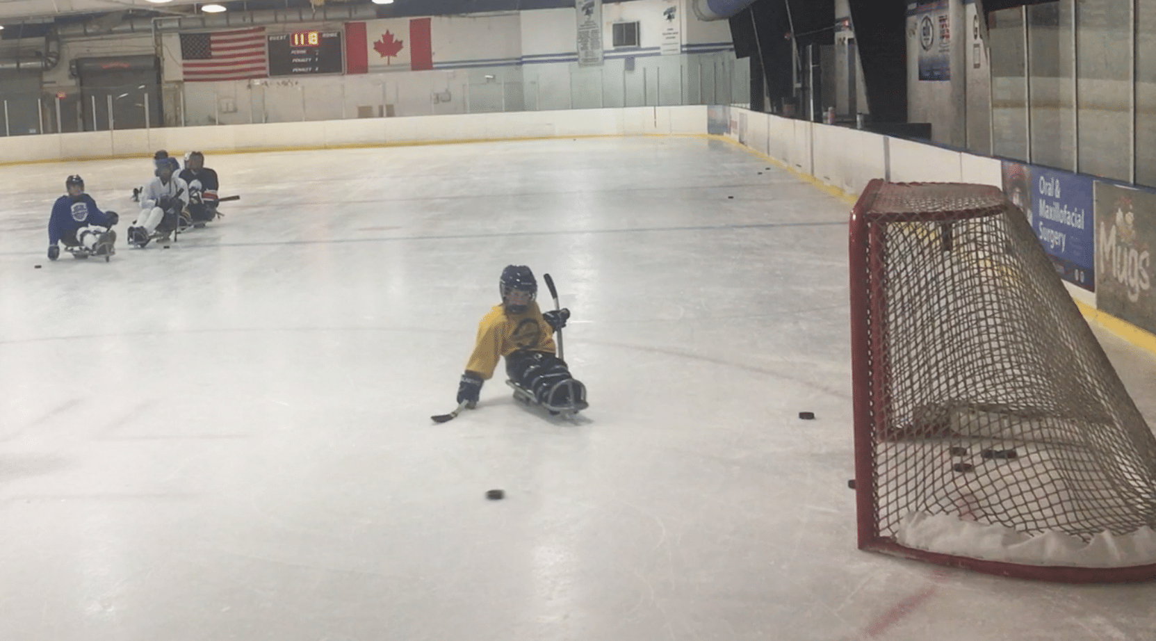 Charlie at the ice rink wearing a yellow jersey and sitting in a hockey sled. He is about to strike a puck toward the goal.