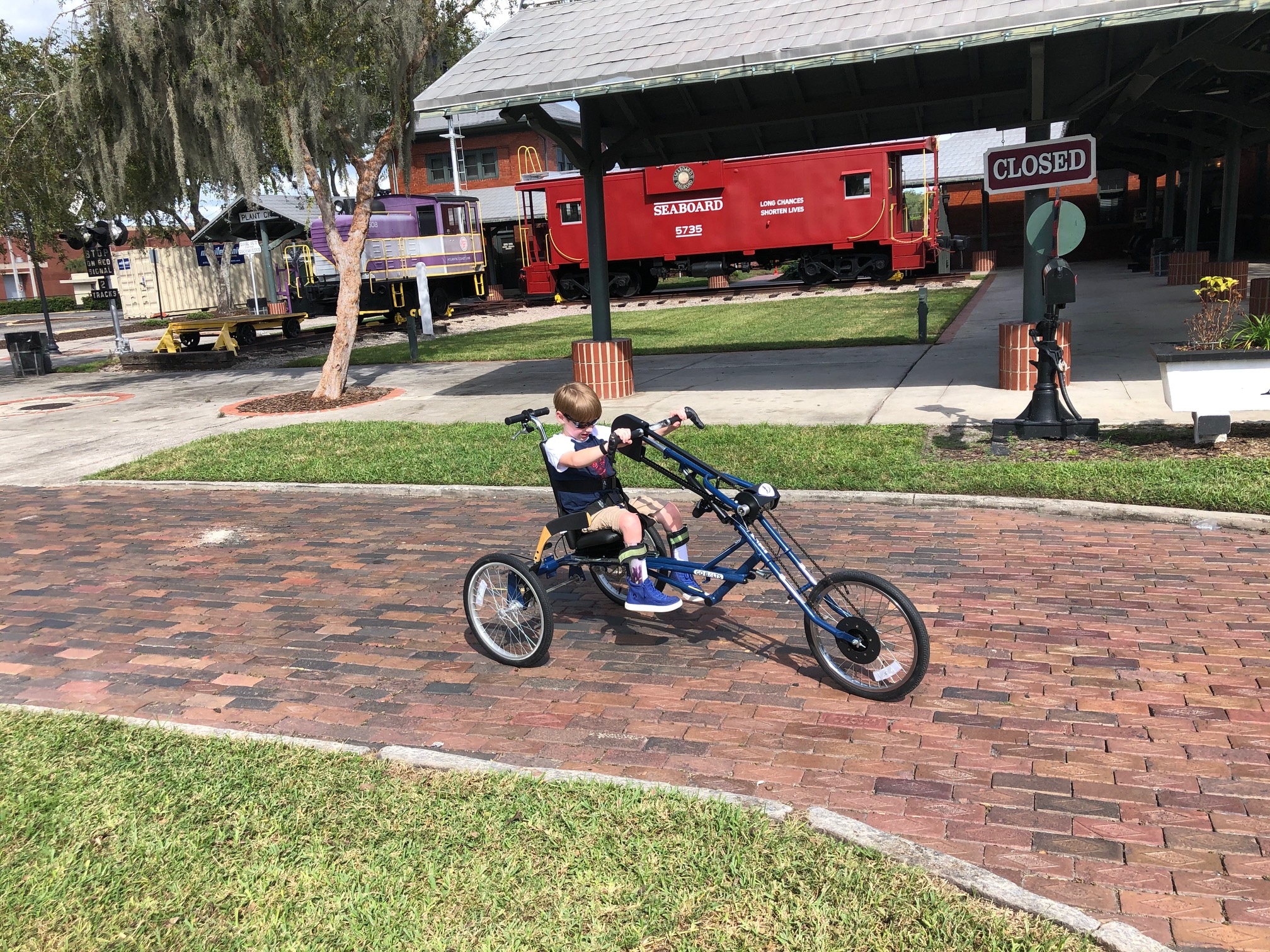 A 9-year-old boy on his three-wheeled handcycle riding down a brick sidewalk with a train caboose in the background.