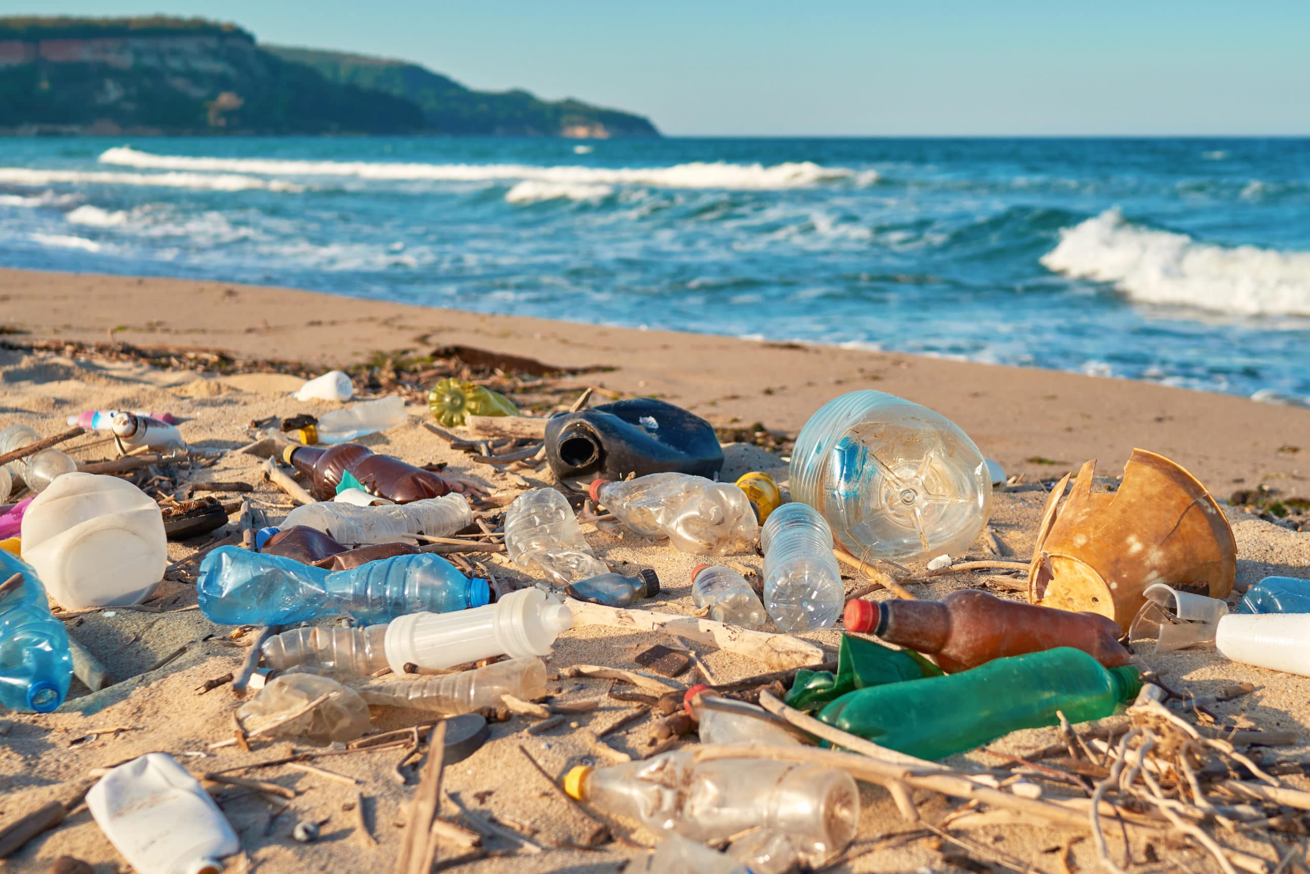 Garbage on a sandy beach with moving waves and a green hill in the background.