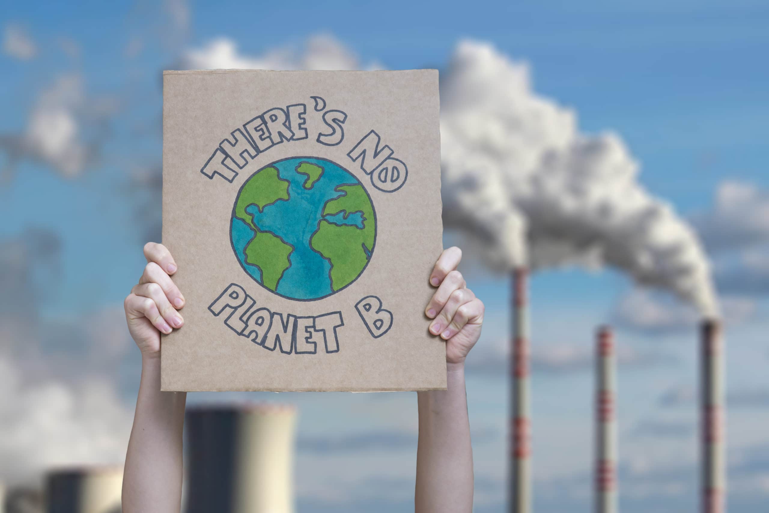 Two hands hold a cardboard poster that reads, "There's no Planet B" with industrial fossil fuel towers spewing smoke in the background.