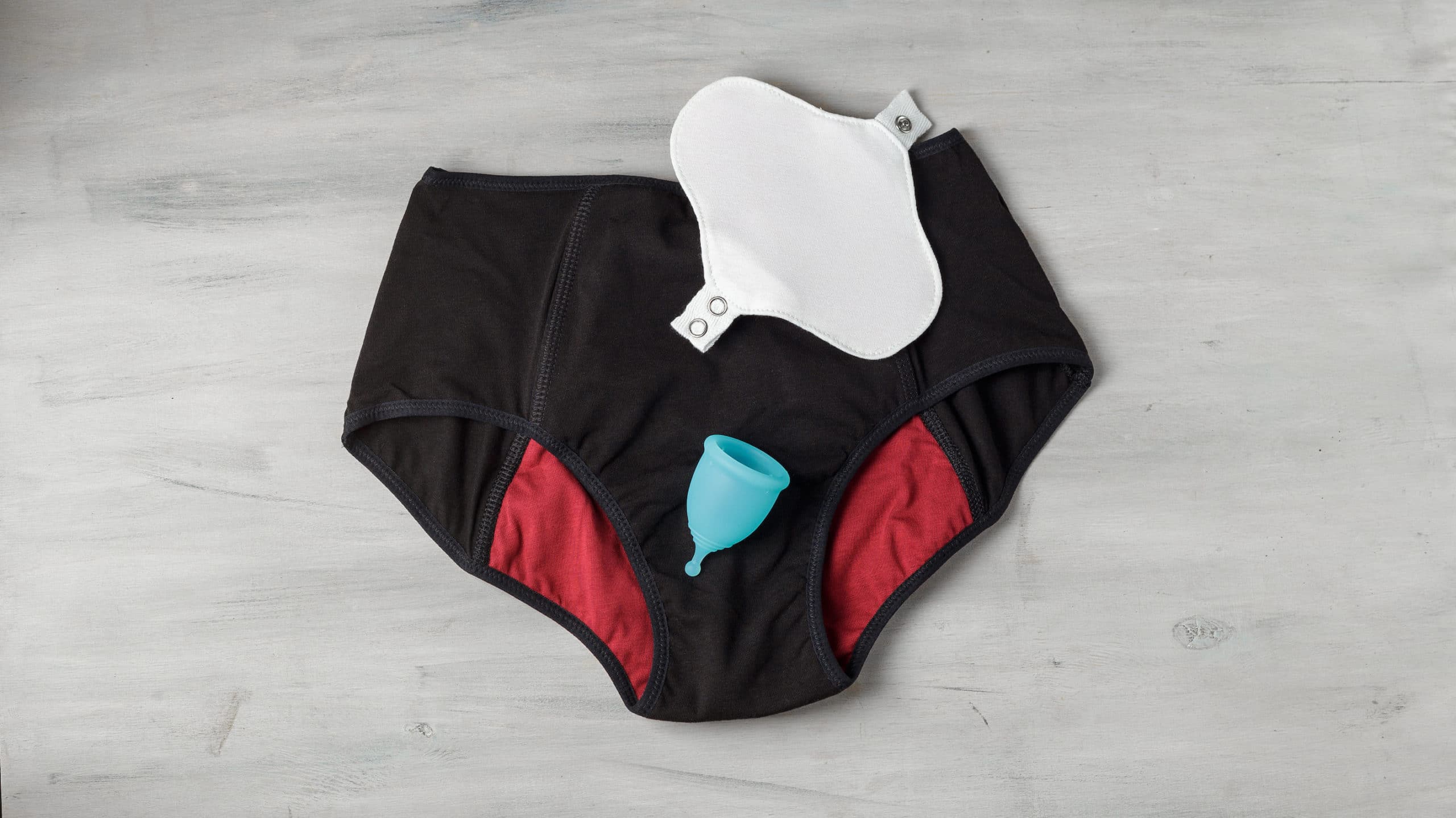 Reusable eco-friendly feminine hygiene period products - menstrual cup and black menstruation pants and organic cotton cloth everyday pad. Top view on grey background.