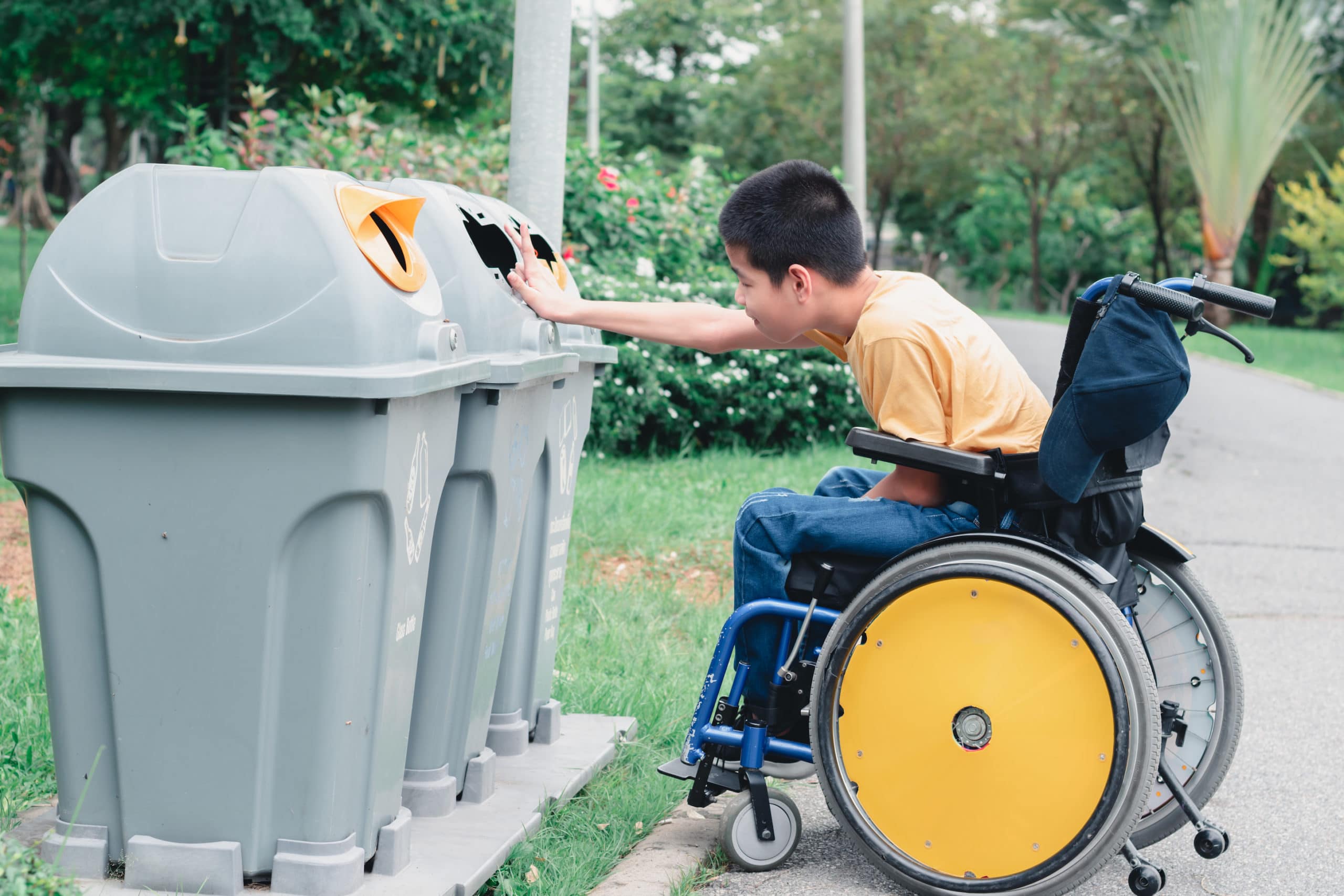 Young boy in wheelchair with yellow wheels reaches from his chair to put an item in one of three recycling or trash bins.