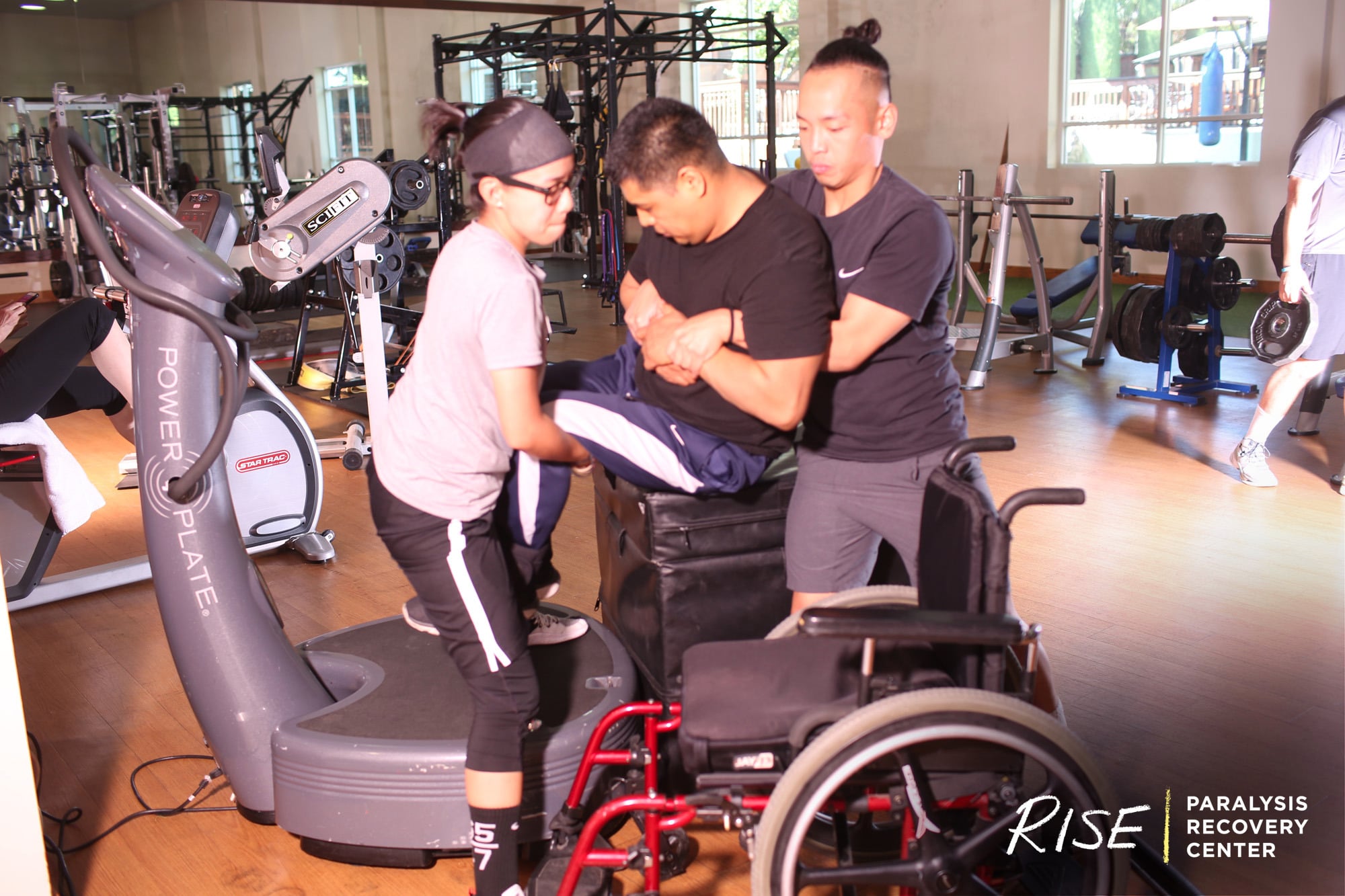 Two trainers help a man get out of his wheelchair onto a machine at the gym.