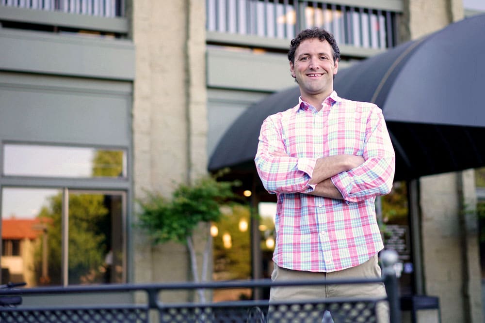 Robert Jackson smiles at the camera with is arms crossed. He is wearing a pink plaid button down shirt.