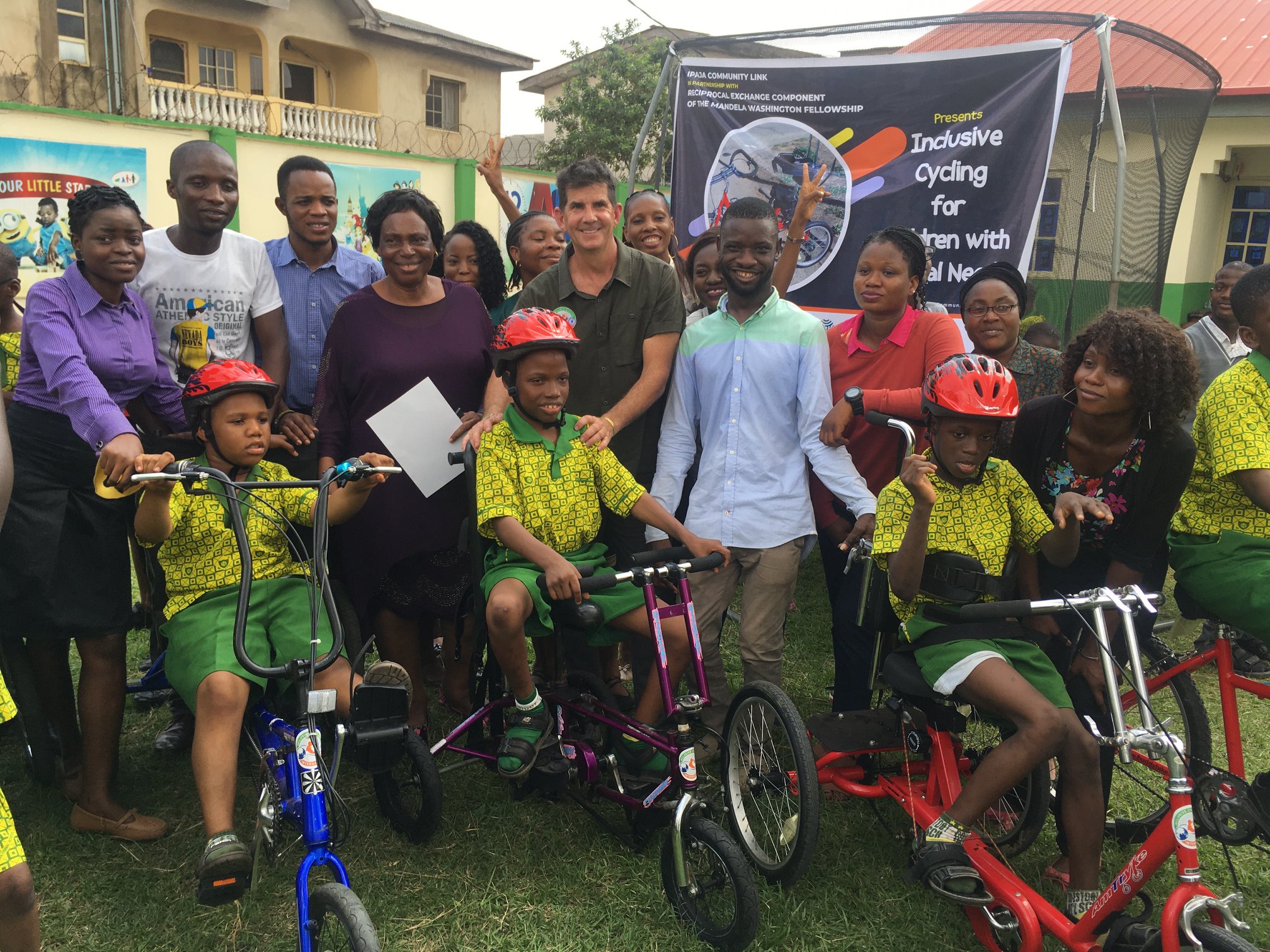 A group of adults stand behind three children on adaptive bicycles. An Inclusive Cycling banner is behind them.