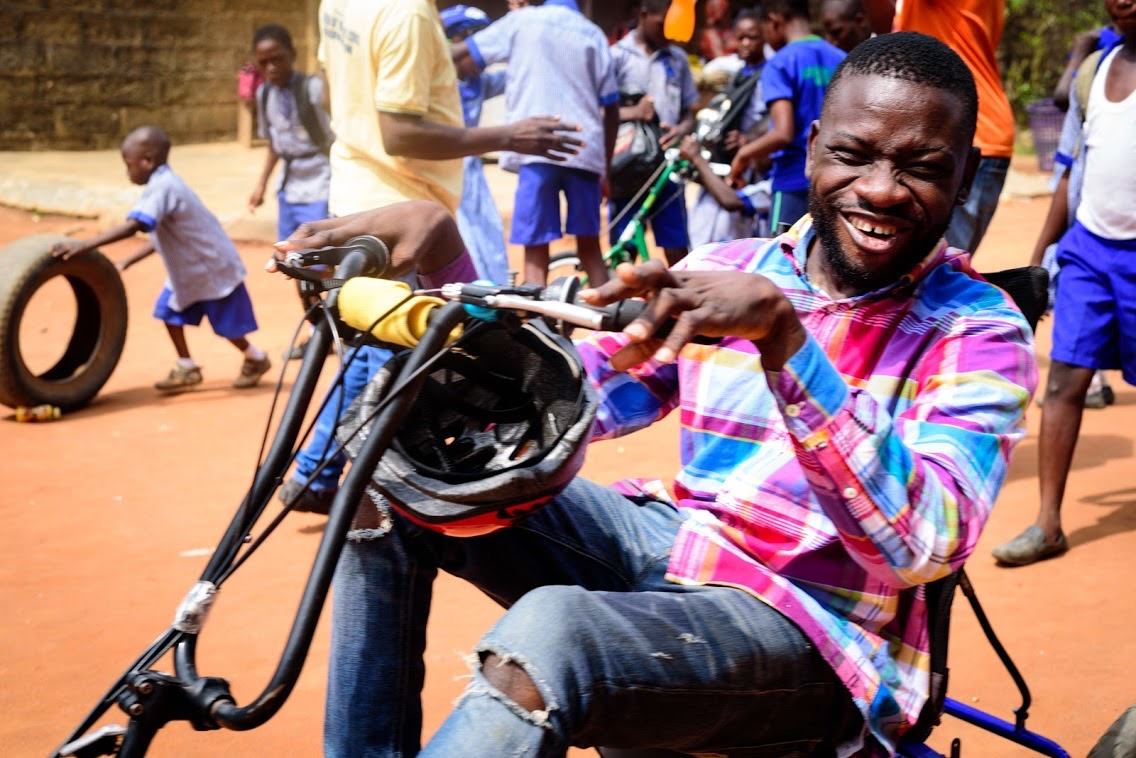 A young Nigerian man smiles big while sitting on an adaptive bike.