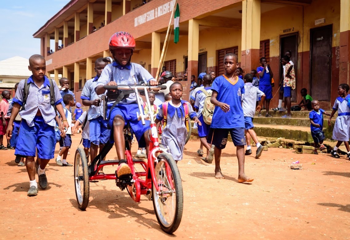 A young Nigerian boy pedals an adaptive bike while his classmates walk alongside him. 