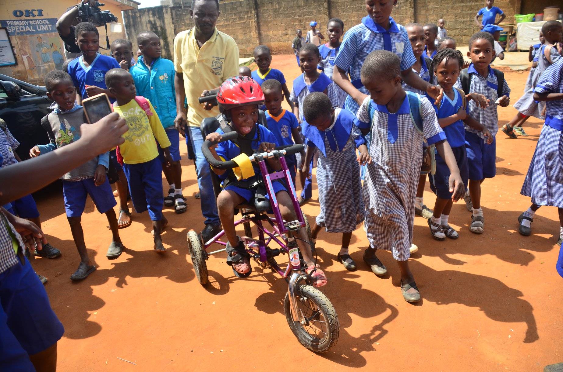 A young Nigerian boy smiles with awe as he pedals an adaptive bike. His classmates and some adults surround him.