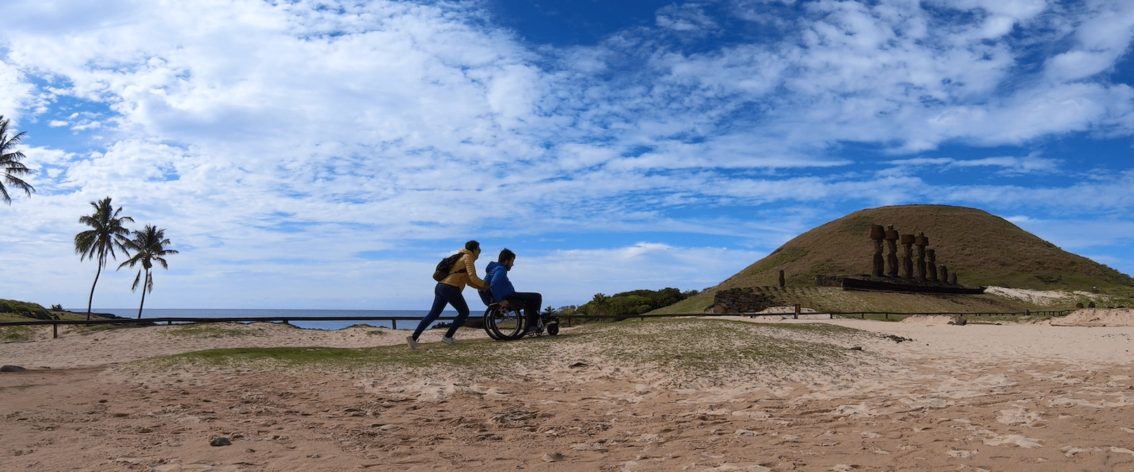 Myriam pushes Pierre in his wheelchair across a sandy, tropical island.