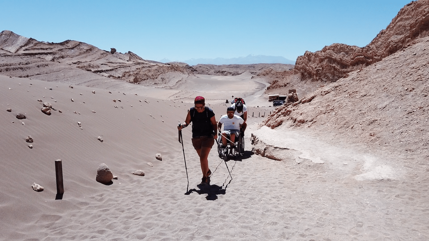 Myriam has walking sticks and ropes attached to Pierre and his chair behind her. Another man helps push Pierre in his chair up a sandy path in the Valley of the Moon in Chile.
