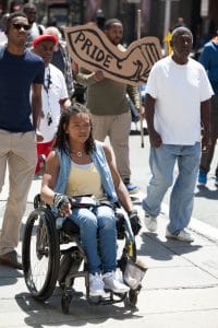 A woman in a wheelchair at the Disability Pride Philadelphia parade. Several people are marching behind her. One holds a "Pride" sign in the shape of a forearm and fist.