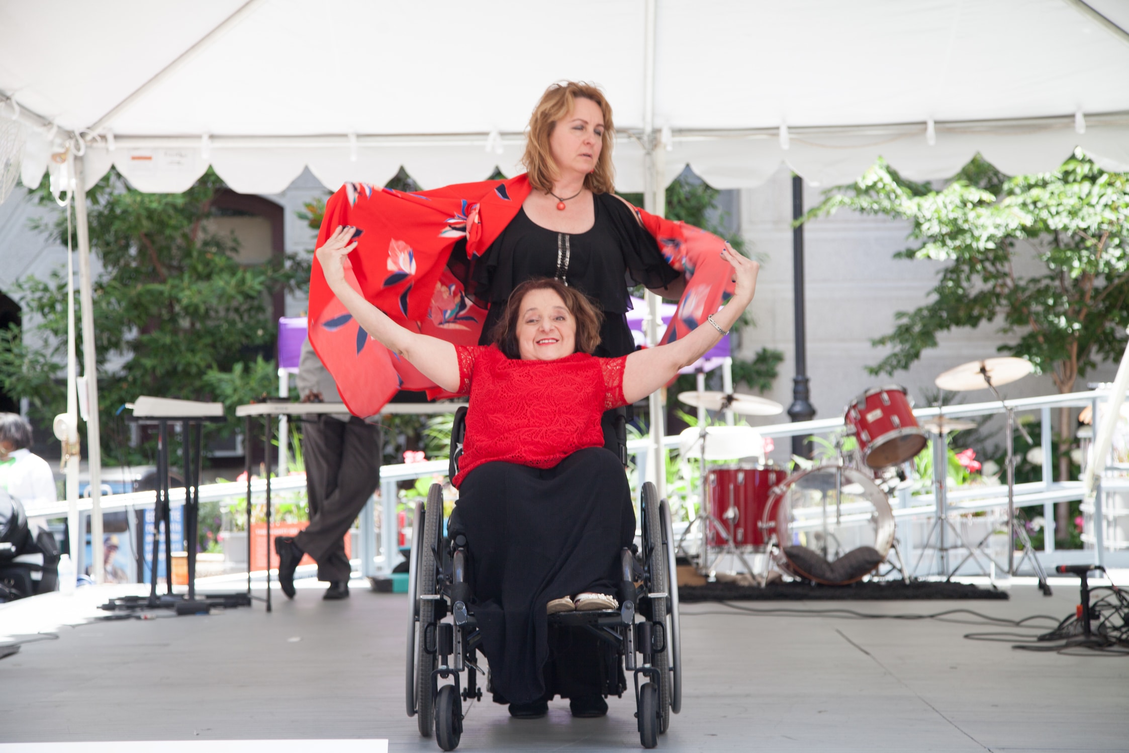 Two women wearing red and black on a stage. One woman is in a wheelchair holding her arms out wide while the other woman does the same while standing behind her.