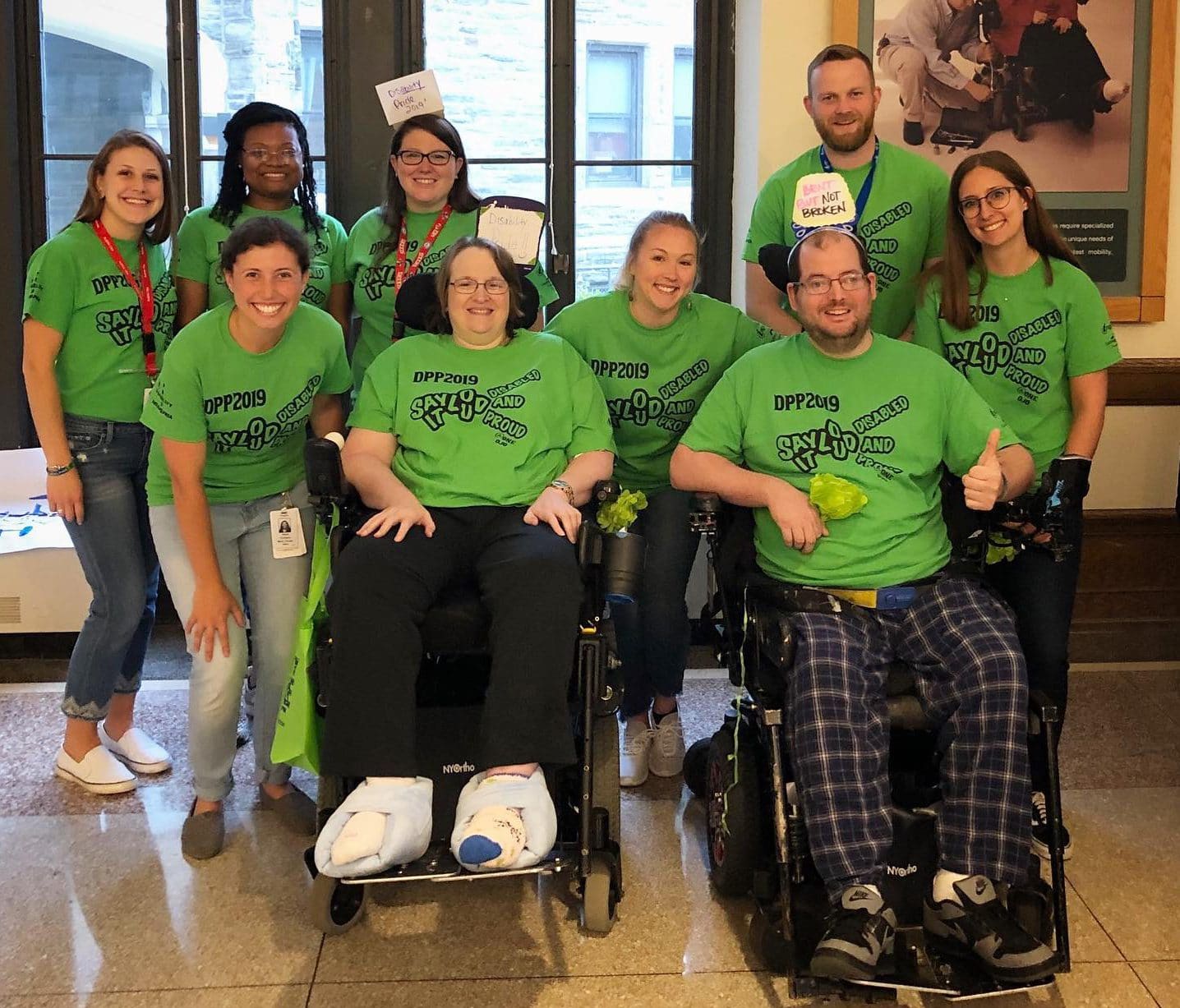 A group of people standing and in wheelchairs wearing green "Say it Loud, Disabled and Proud" t-shirts.