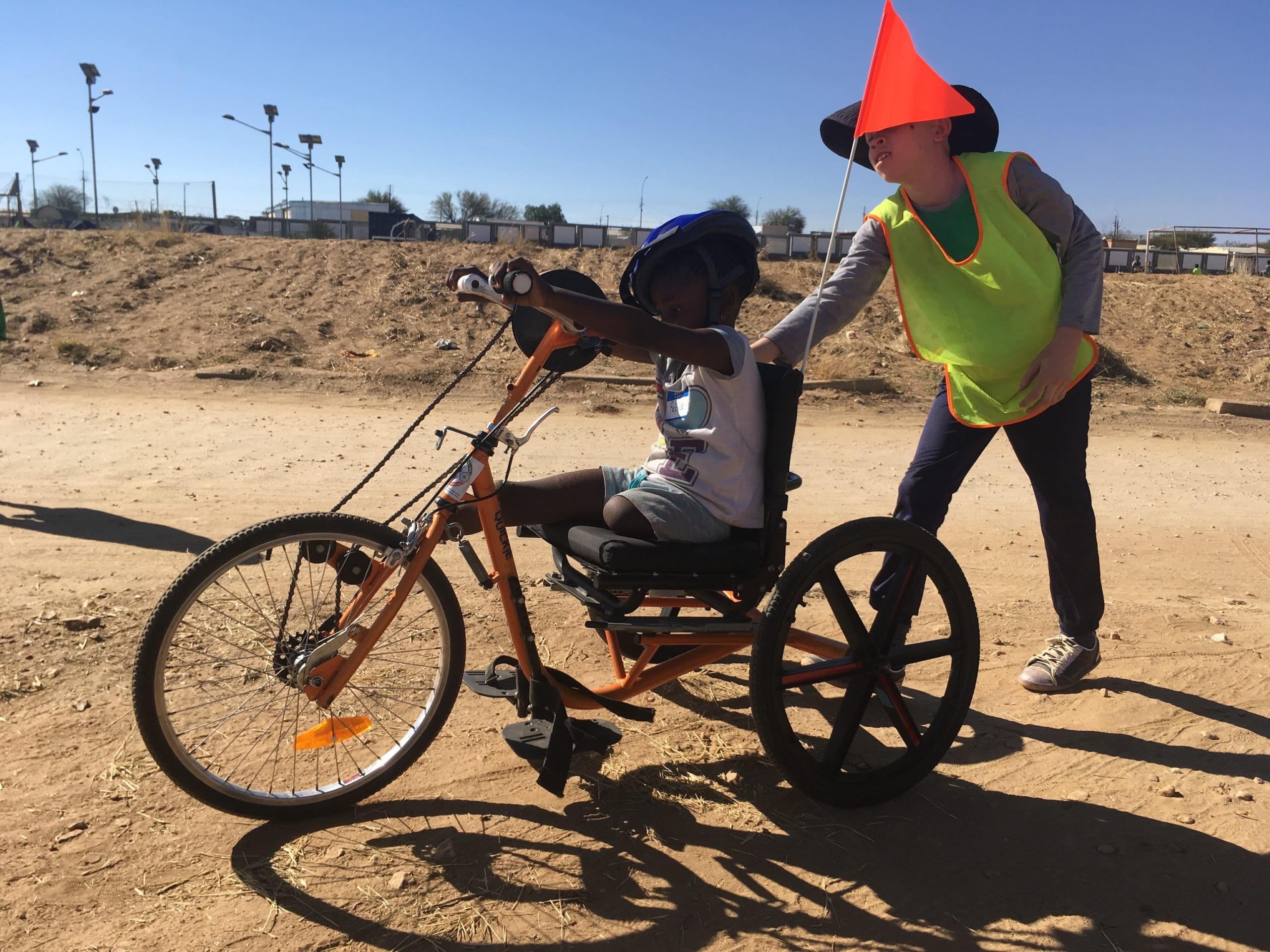 A leg-amputee child sits on an adaptive bike while another child stands behind with one hand on the bike as if he's pushing it.